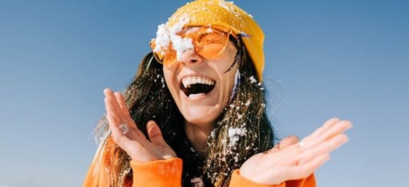 A young woman wearing sunglasses is laughing and playing with snow.