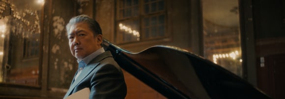 Chinese classical music conductor Long Yu stands in front of a piano with sheet music in his hand.