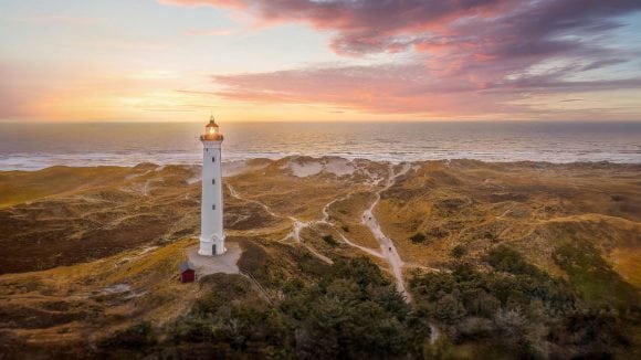 White lighthouse standing among coastal dunes overlooking the sea at sunset