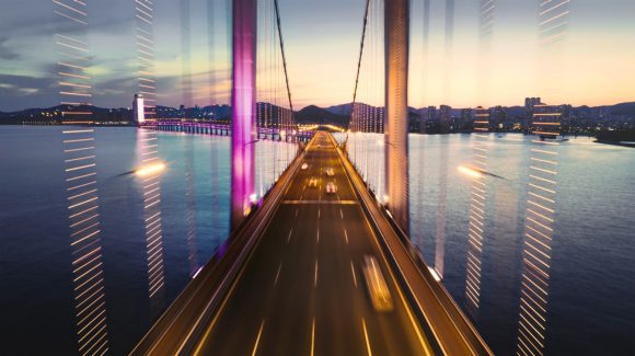 A modern bridge illuminated at dusk, with light trails suggesting motion, above water and a city skyline in the background.