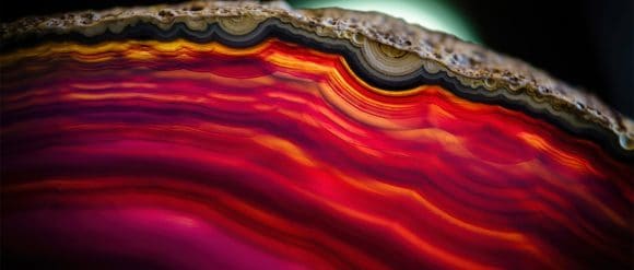 Abstract close-up of layered, agate-like bands in vivid red, orange, and pink, with a dark edge and soft glow in the background.