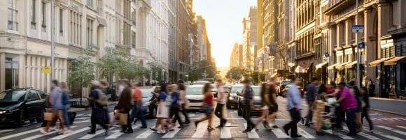 Crowd of blurred, fast-paced, city-dwellers walking across a crosswalk while the urban environment behind them stands.