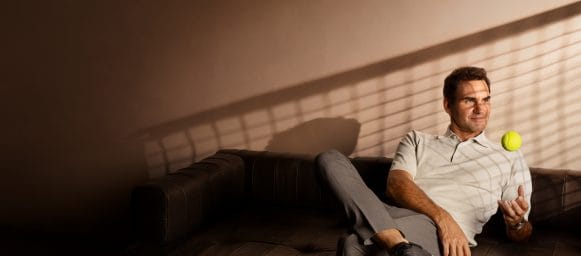 El tenista Roger Federer está sentado en un sofá con una pelota de tenis amarilla en la mano. La sombra de una red de pista de tenis se refleja en la pared detrás de él.