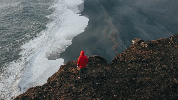 Aerial view of a figure in a red coat standing on a cliff overlooking waves washing up on a dark sandy beach.
