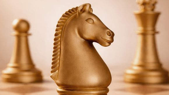Close up view of a gold-hued chess board featuring the knight (horse) in the foreground and a pawn to the left and the queen to the right in the background.