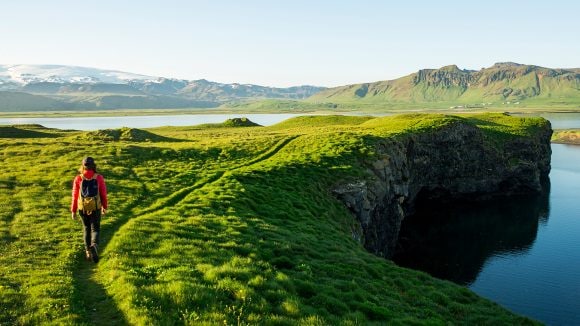 Image of a hiker in a red coat and green backpack walking along a trail in a rich green terrain toward a lake with mountains along the horizon.
