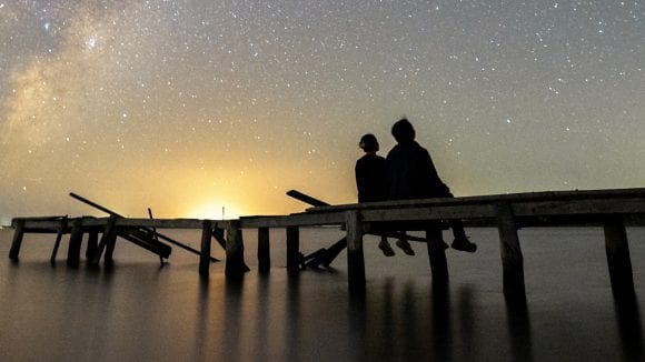 A couple siting on a long dock seen from behind who are gazing at the sunset.