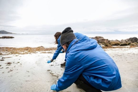 Eco-friendly female scientists gathering micro plastic specimens on rugged beach.