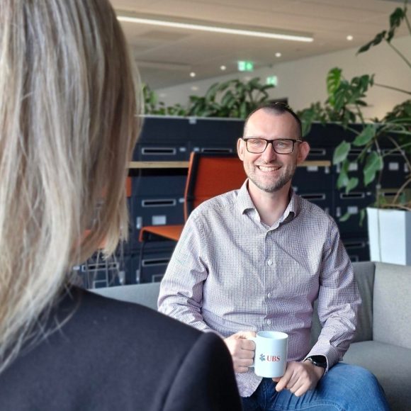 Wojtek chats with a teammate over coffee on a couch in the office. He smiles warmly. Flowers and a cosy workspace appear behind them.