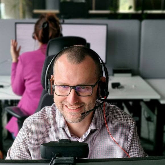 Wojtek sits at his desk in an online meeting, smiling and focused with a headset on. The open office and colleagues are visible behind him.