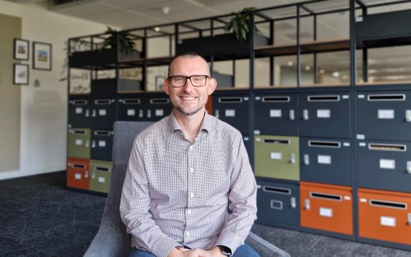 Wojciech sits smiling in an armchair in the office meeting area. He wears a plaid shirt and glasses. Flowers and colourful shelves are visible in the background.