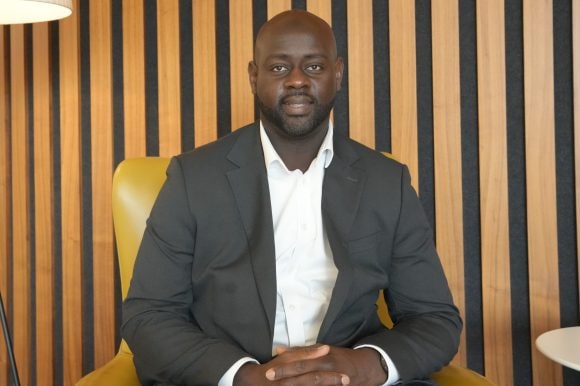 Abdul sits upright in a yellow armchair against a backdrop of vertical wooden stripes. He wears a black suit with a white shirt, looking directly at the camera.