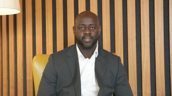 Abdul sits upright in a yellow armchair against a backdrop of vertical wooden stripes. He wears a black suit with a white shirt, looking directly at the camera.