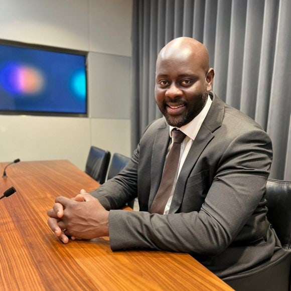 Abdul sits at a wooden conference table, dressed in a gray suit with a white shirt and a brown tie. He is resting his hands on the table in front of him. The background features a muted gray curtain and a wall-mounted screen.