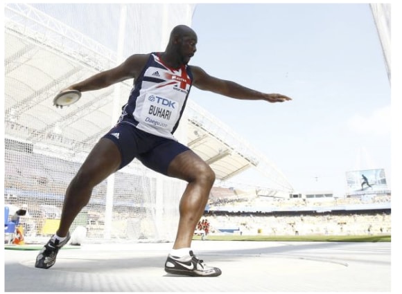 Abdul prepares to throw a discus at the London 2012 Olympic Games. He wears a team GB athletic uniform with 'Buhari' on the name tag. The stadium is filled with spectators in the background.