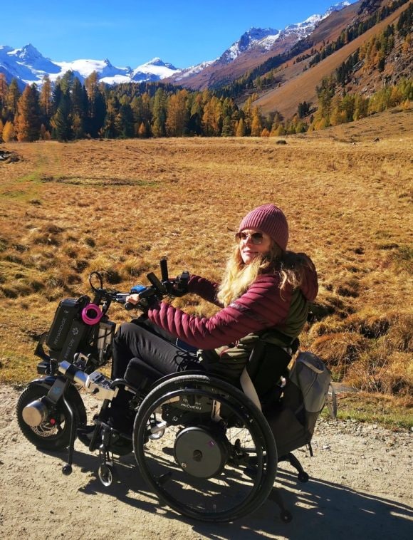 The image shows Samira sitting in her off-road wheelchair and experiencing the beauty of the Swiss mountains in a golden autumn scenery.