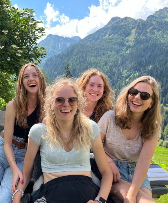 The image shows Samira sitting outdoors with close friends during a trip to the Swiss mountains, smiling at the camera amid lush green spring scenery.