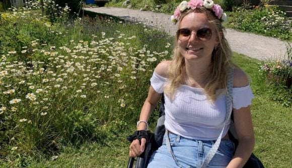 The image shows Samira sitting in a wheelchair in a green spring scenery, smiling at the camera. She wears sunglasses and a flower wreath.