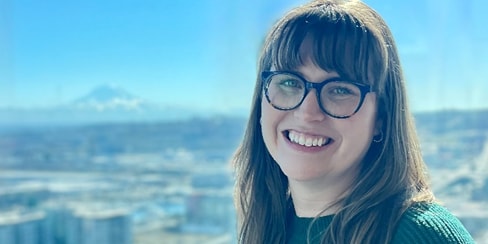 Woman wearing glasses standing in front of a scenic view that has blues and greens. She's smiling and wearing a green sweater