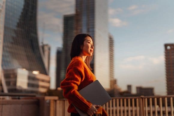 Una mujer sujetando su computadora mirando la vista de una ciudad