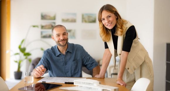 Dos personas en oficina sonríen mientras revisan planos y maqueta arquitectónica en una mesa.