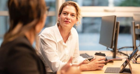 Mujer en oficina conversando con colega frente a computadora y taza de café.