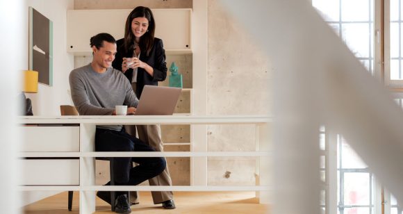 Dos personas conversan en un espacio interior moderno con barandal y ventanales.