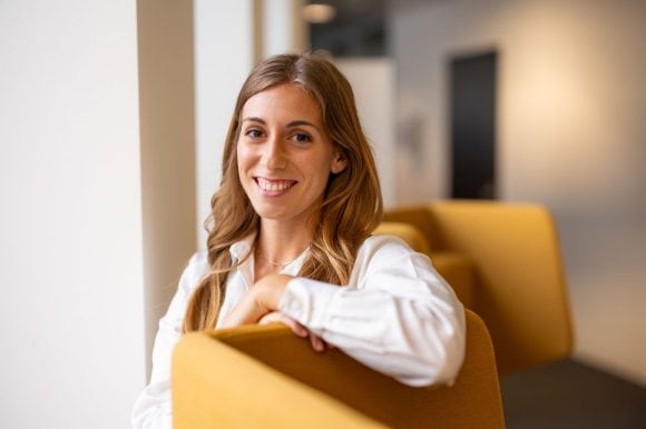 Mujer sonriente apoyada en respaldo de silla en entorno de oficina.
