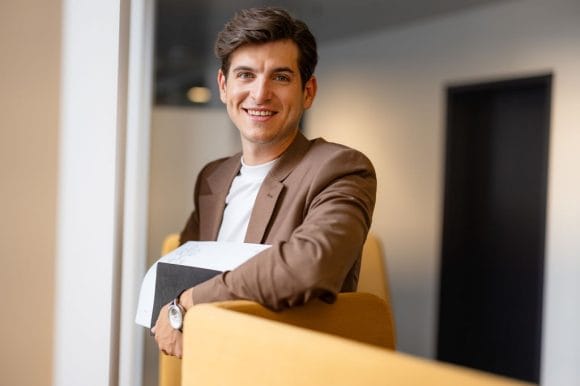 Hombre sonriente apoyado en respaldo de silla en un espacio interior.