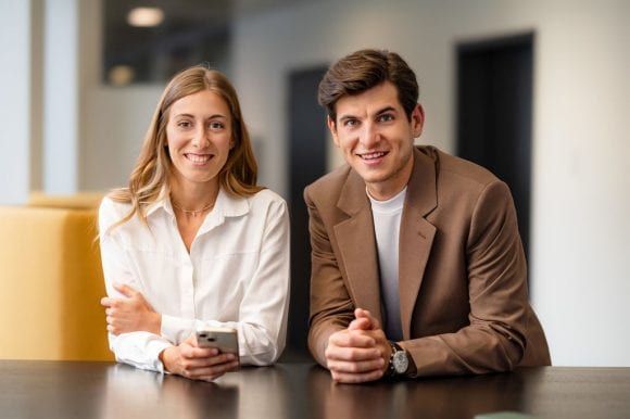 Dos personas sonríen a cámara sentadas en una mesa en entorno de oficina.