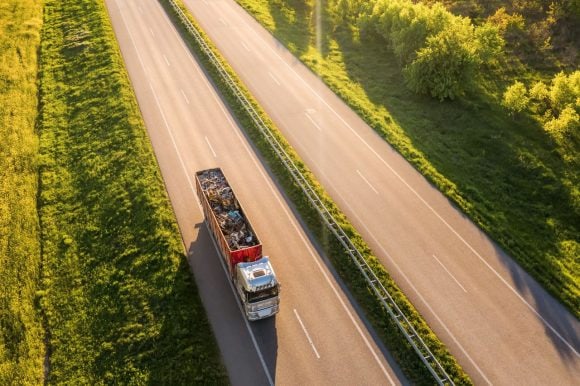 Camión de carga transportando residuos por carretera rodeada de vegetación.