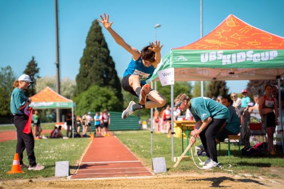 Ein dunkelhaariger Junge (ca. 12) springt beim Weitsprung auf dem Sportplatz, ein Fuss nach vorne, Arme oben. Zwei Erwachsene beobachten den Absprung. Im Hintergrund grüne Bäume, blauer Himmel, zwei UBS Kids Cup Pavillons (grün/orange) und wartende Kinder.