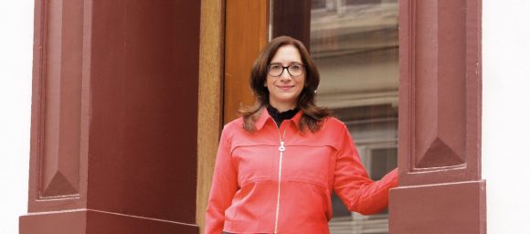 Braunhaarige Frau mit Brille und rotem Oberteil mit weissem Reissverschluss steht vor einer Türe und platziert ihre linke Hand locker an der Wand.