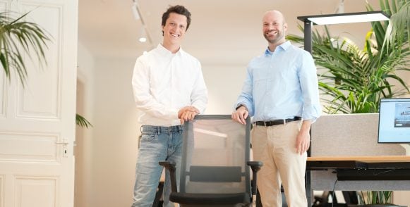 Two smiling men stand behind a desk chair in an office.