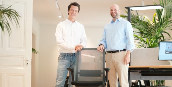 Deux hommes souriants se tiennent debout derrière un fauteuil de bureau.