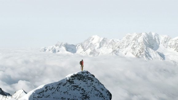 Um montanhista no pico de uma montanha nevada com vista para um vale coberto de nuvens. 
