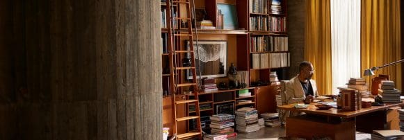 Hombre sentado en un escritorio en una oficina iluminada por la luz del sol, leyendo un libro, rodeado de estanterías y pilas de libros.