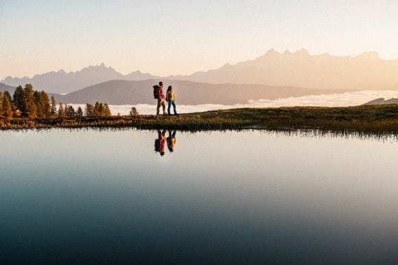 Man and woman hiking along a lake with mountains in the background