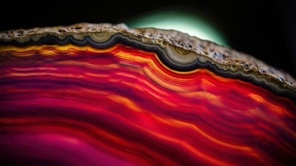 Abstract close-up of layered, agate-like bands in vivid red, orange and pink, with a dark edge and soft glow in the background