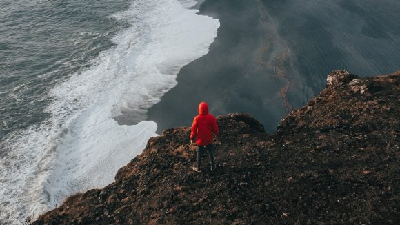 Aerial view of a figure in a red coat standing on a cliff overlooking waves washing up on a dark sandy beach.