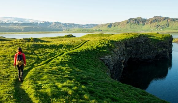 Image of a hiker in a red coat and green backpack walking along a trail in a rich green terrain toward a lake with mountains along the horizon.