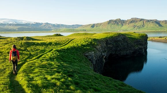 Image of a hiker in a red coat and green backpack walking along a trail in a rich green terrain toward a lake with mountains along the horizon.