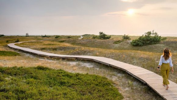 Woman with a hat walking barefoot along a boardwalk across low dunes toward the ocean with sun shining from beneath clouds.