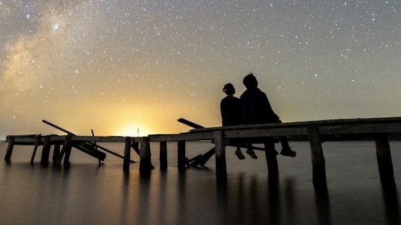 A couple siiting on a long dock seen from behind who are gazing at the sunset.