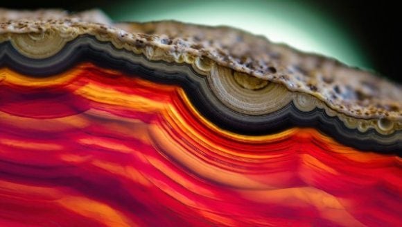 Abstract close-up of layered, agate-like bands in vivid red, orange and pink, with a dark edge and soft glow in the background