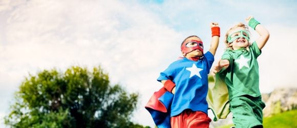 Children dressed as superheroes in capes and masks smiling outdoors.
