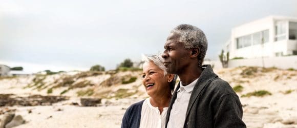 Senior couple walking arm in arm on the beach, home in background