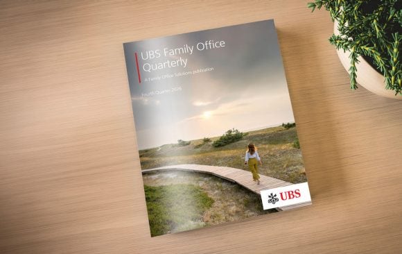 Photograph of a UBS Family Office Quarterly report cover placed on a wooden desk. Cover shows a coastal landscape with a person on a curved boardwalk, “Fourth Quarter 2024” text, and UBS logo at bottom right.