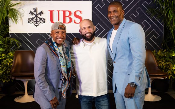 Kevin Liles, Steve Rifkind and Wale Ogunleye grouped together, smiling at the camera with UBS logo in the background