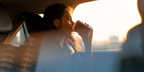 A young Asian woman sips a coffee in the back seat of a car, taking in the view at sunrise or sunset